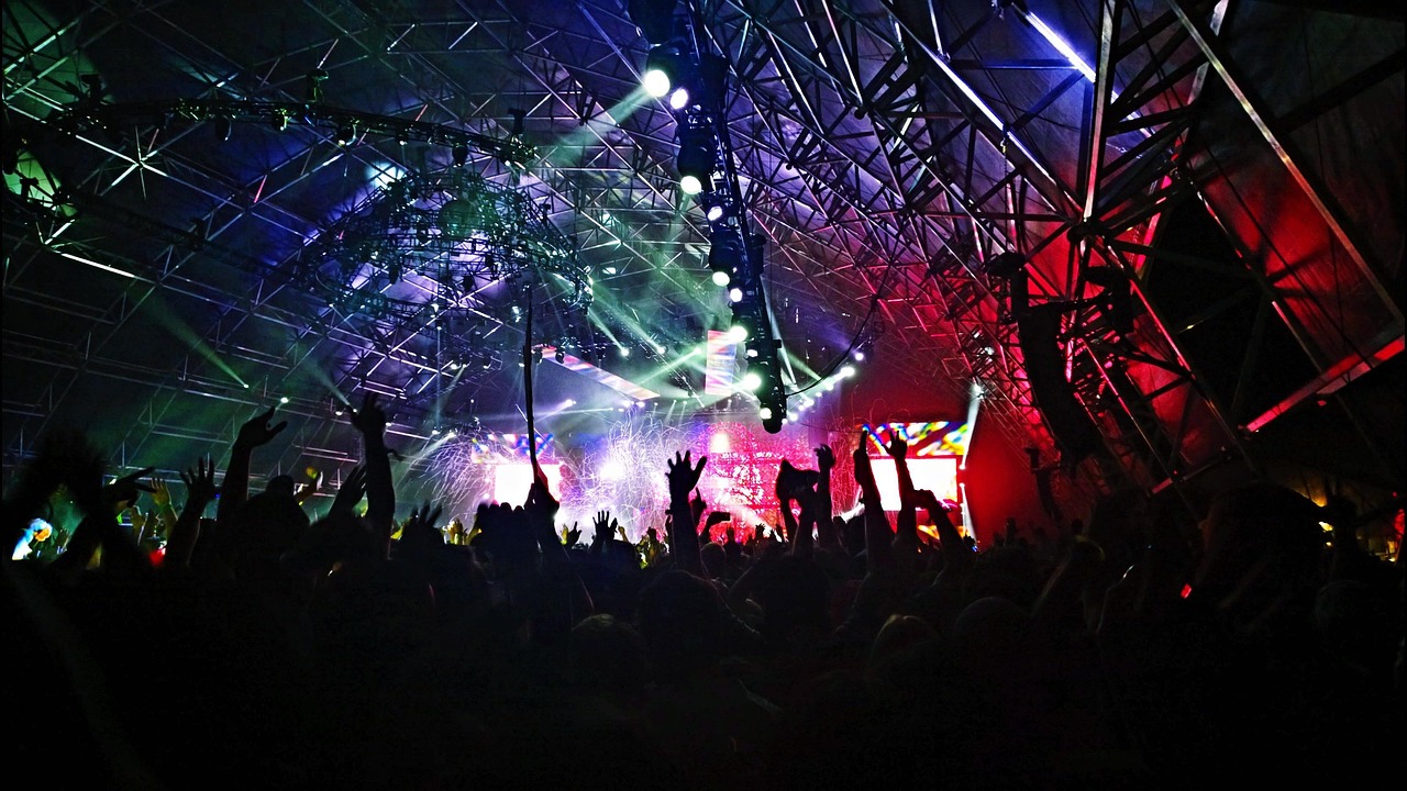 A large crowd of fans with hands raised, illuminated by colorful stage lights at a live concert.